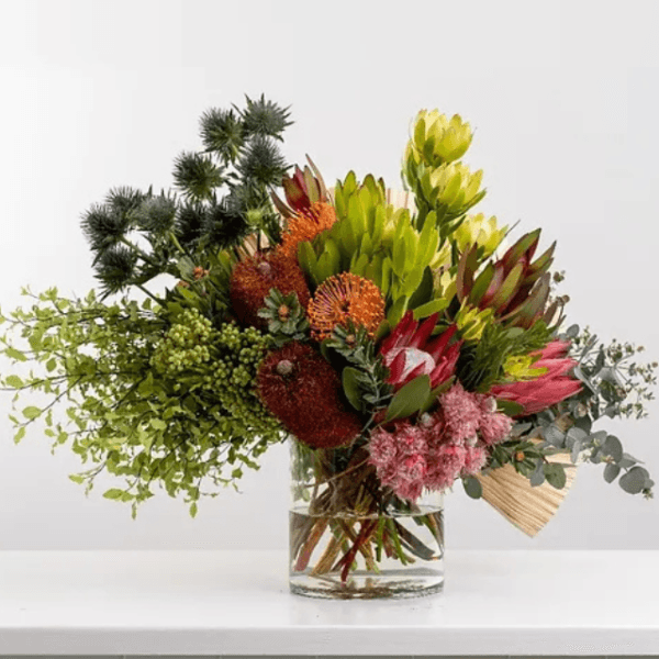 Rustic Autumn Bloom Vase with protea, thistle, and greenery in a clear glass vase, styled on a wooden table with autumn leaves.
